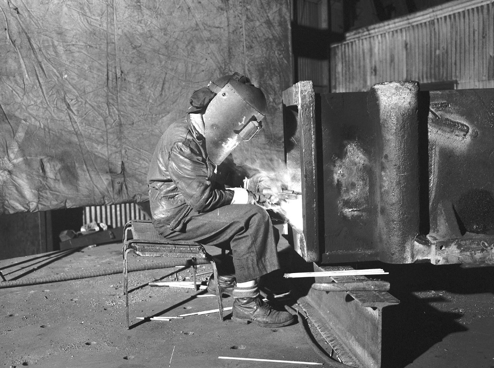 A woman sits on a stool wearing a protective mask while welding a metal beam