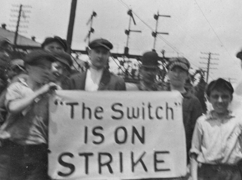 Men and boys stand around a sign reading “’The Switch’ is on strike”