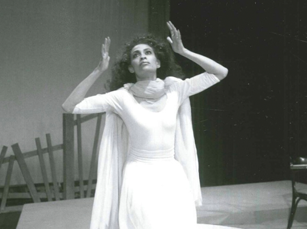 A woman dressed in white kneels on a pedestal on a stage and raises her hands to frame her head
