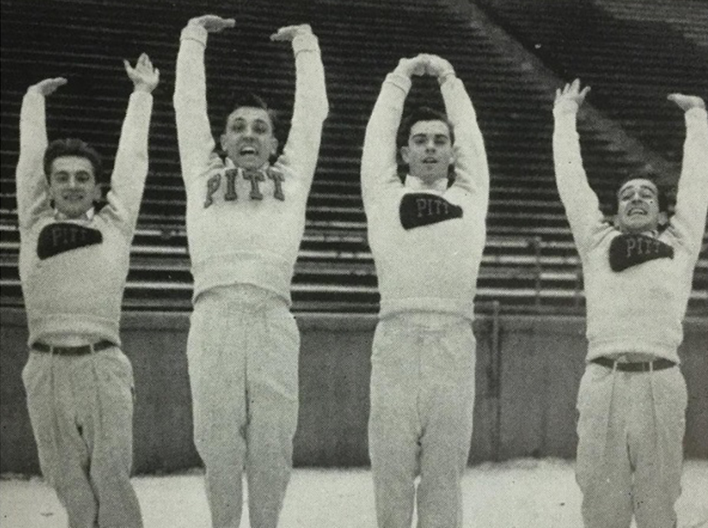 Four male cheerleaders wearing Pitt sweaters jump with their arms raised