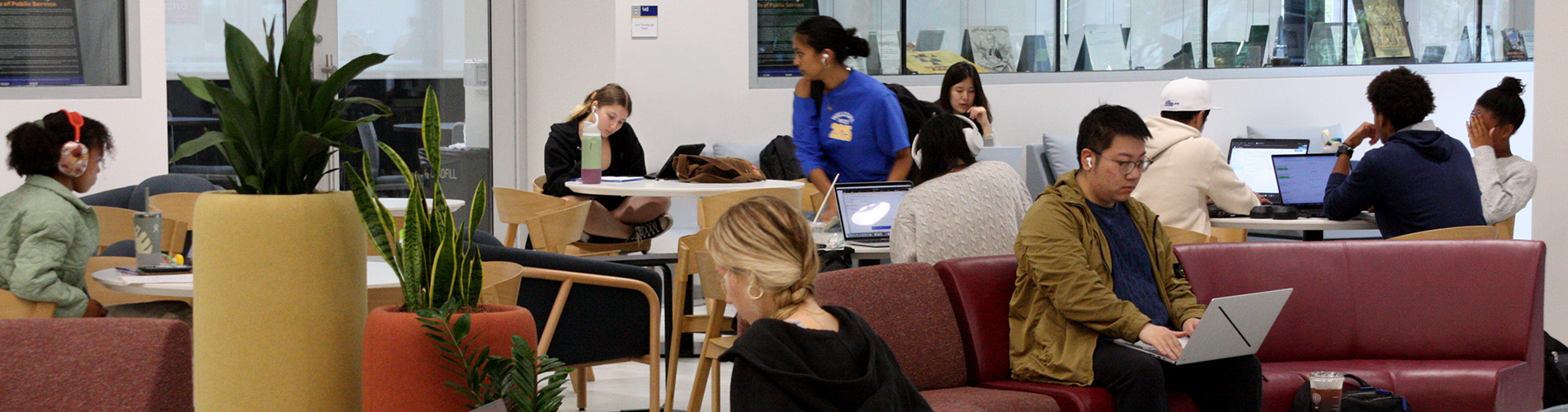 People studying on the first floor of the University of Pittsburgh Hillman Library during an open house event. Several individuals are seated at tables and couches, using laptops and study materials. Plants are in the foreground, and a glass wall with bookshelves is visible in the background.