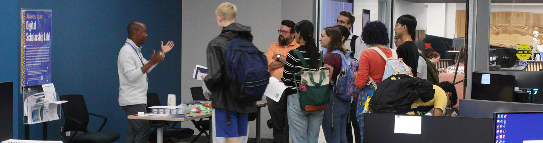 A group of people gathered in the Digital Scholarship Lab at the University of Pittsburgh Hillman Library during an open house. They are listening to a presenter explaining the space, with computer stations and informational posters visible in the background. Some attendees carry backpacks and hold notebooks or pamphlets.