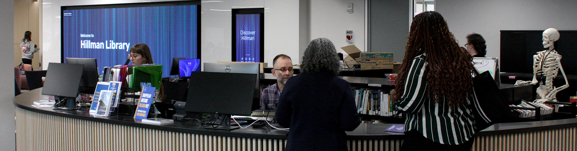 Visitors getting help at the Donald Steele Wood Service Desk during the Hillman Library open house. The desk has monitors, brochures, and a skeleton model. A screen behind says 'Welcome to Hillman Library.
