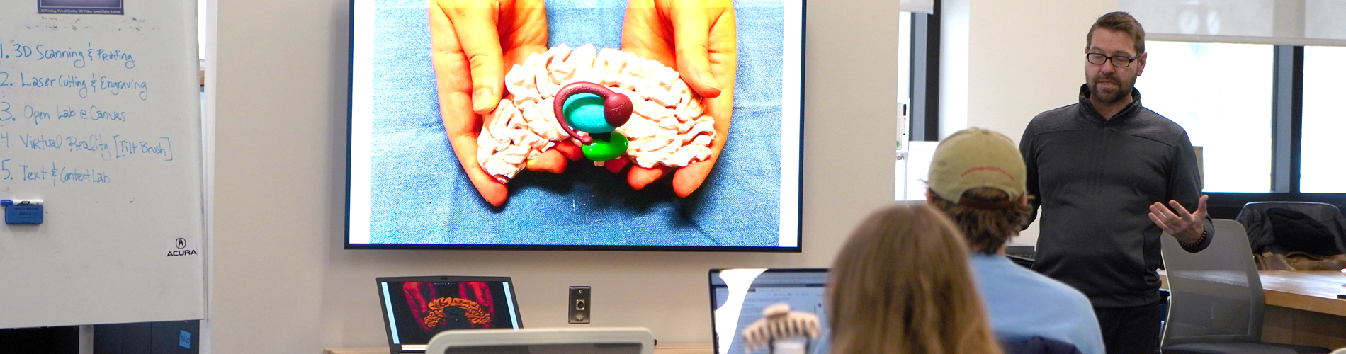 An instructor in the Open Lab gives a lecture to students, with a large screen showing a 3D printed model of the human brain. A whiteboard nearby lists lab activities such as 3D scanning, laser cutting, virtual reality, and text and context lab.