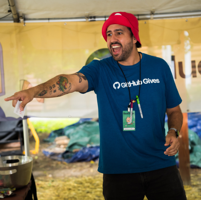 PJ Metz, wearing a red hat and blue GitHub Gives shirt, points toward something off-camera while smiling.