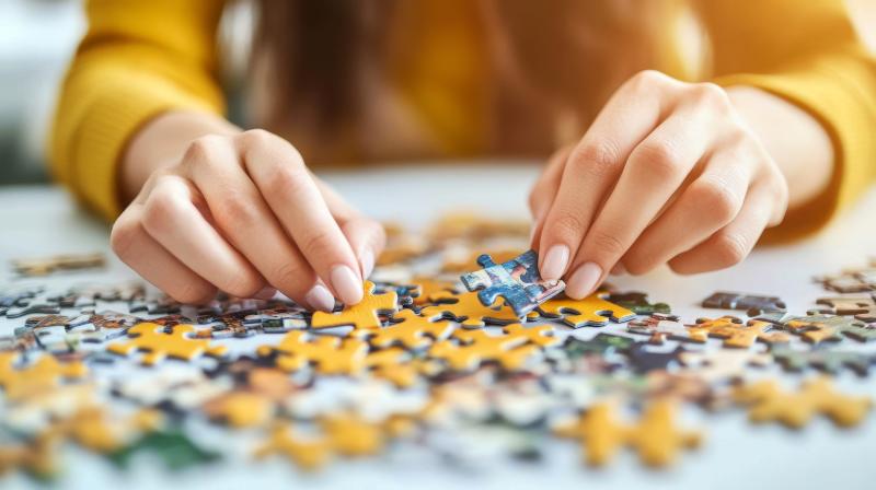 Close-up of a person wearing a yellow sweater placing a blue jigsaw puzzle piece among scattered yellow and multicolored pieces on a table.