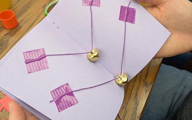 Close-up of hands holding a handmade paper book opened to a page with purple yarn stitched through square holes, connecting to small gold bells attached to the page. The book rests on a wooden table with craft supplies visible in the background