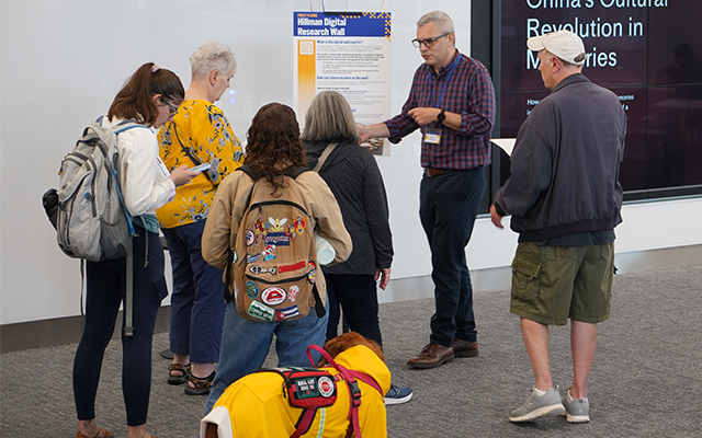 A librarian leads a small group of people on a library tour, standing in front of a digital research wall display while explaining resources.